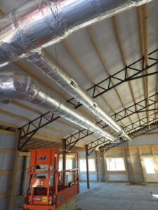 A construction site interior with exposed insulated ductwork on the ceiling, metal trusses, wooden framing, and a scissor lift positioned below.