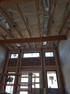 Exposed ceiling with insulation and ductwork in a room under construction, featuring large windows and unfinished walls.