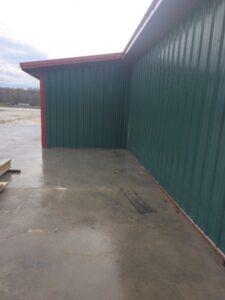 A concrete slab next to a green metal building with red trim, photographed outdoors on a cloudy day. Some wooden boards and tire marks are visible on the ground.