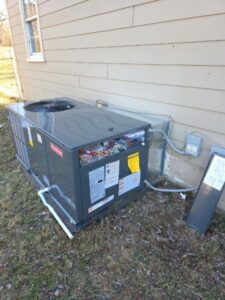 A large HVAC unit sits on the ground outside a beige house, with its side panel open exposing internal wiring; nearby, a gray electrical box is open.