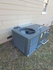 A gray outdoor air conditioning unit is installed on the ground next to the exterior wall of a house.