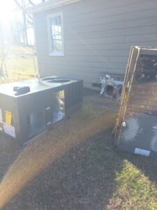 A large HVAC unit sits on the ground outside a house, with a removed metal panel nearby and a vent visible on the house wall.