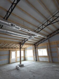 Interior of a partially constructed building with exposed wooden beams, metal supports, large windows, and HVAC ductwork on the ceiling.