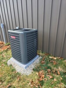 A Goodman outdoor air conditioning unit is installed on a gravel base next to a brown metal-sided building, with some fallen leaves on the grass.