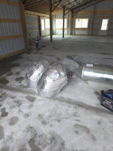 Several large, silver, insulated duct pieces are lying on the concrete floor of a partially constructed building with exposed framing and unfinished walls.