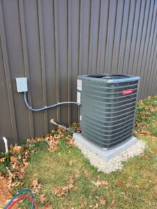 Outdoor air conditioning unit installed next to a metal building, with electrical conduit connected and fallen leaves scattered on the ground.