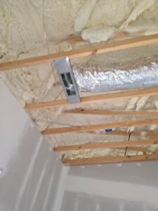 Ceiling under construction with exposed wooden beams, spray foam insulation, ductwork, and partially installed drywall.