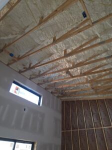 Interior view of a building under construction showing exposed wooden roof trusses, spray foam insulation on the ceiling, and partially installed drywall and insulation on the walls.