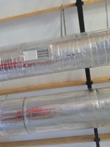 Metal ventilation ducts wrapped in insulation run along a ceiling, with wooden beams and a black metal support visible.
