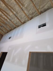 A partially constructed wall with drywall installed and seams taped and mudded, featuring two rectangular wall openings; exposed ceiling beams and spray foam insulation are visible above.