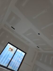 A room with unfinished drywall on the walls and ceiling, showing taped joints and a window partially covered with protective material.