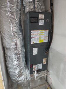 A gray HVAC air handler unit is installed vertically in a utility closet, with insulated ductwork connected on the left and warning labels visible on the front of the unit.