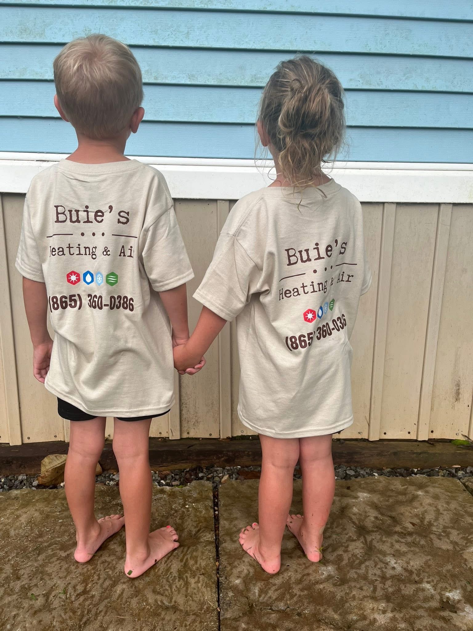 Two children stand side by side holding hands, wearing oversized Buie’s Heating & Air t-shirts, facing a blue and white exterior wall.
