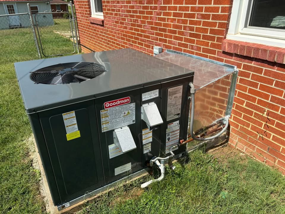 A large Goodman HVAC unit is installed outside a brick house, with metal ductwork connecting it to the building.