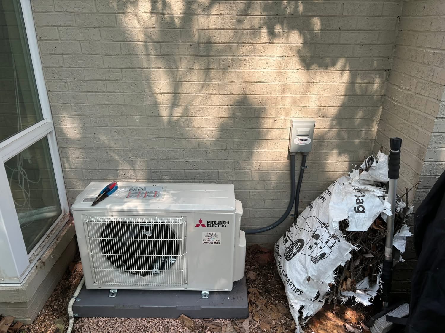 An outdoor Mitsubishi Electric air conditioning unit sits on gravel beside a brick wall, near a pile of bagged leaves and an electrical outlet.