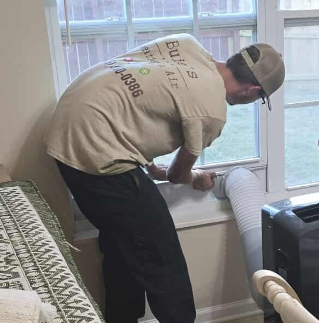 A man in a beige shirt installs a portable air conditioner vent hose into a window inside a room with a patterned couch, ready to handle the heat while awaiting emergency HVAC services.