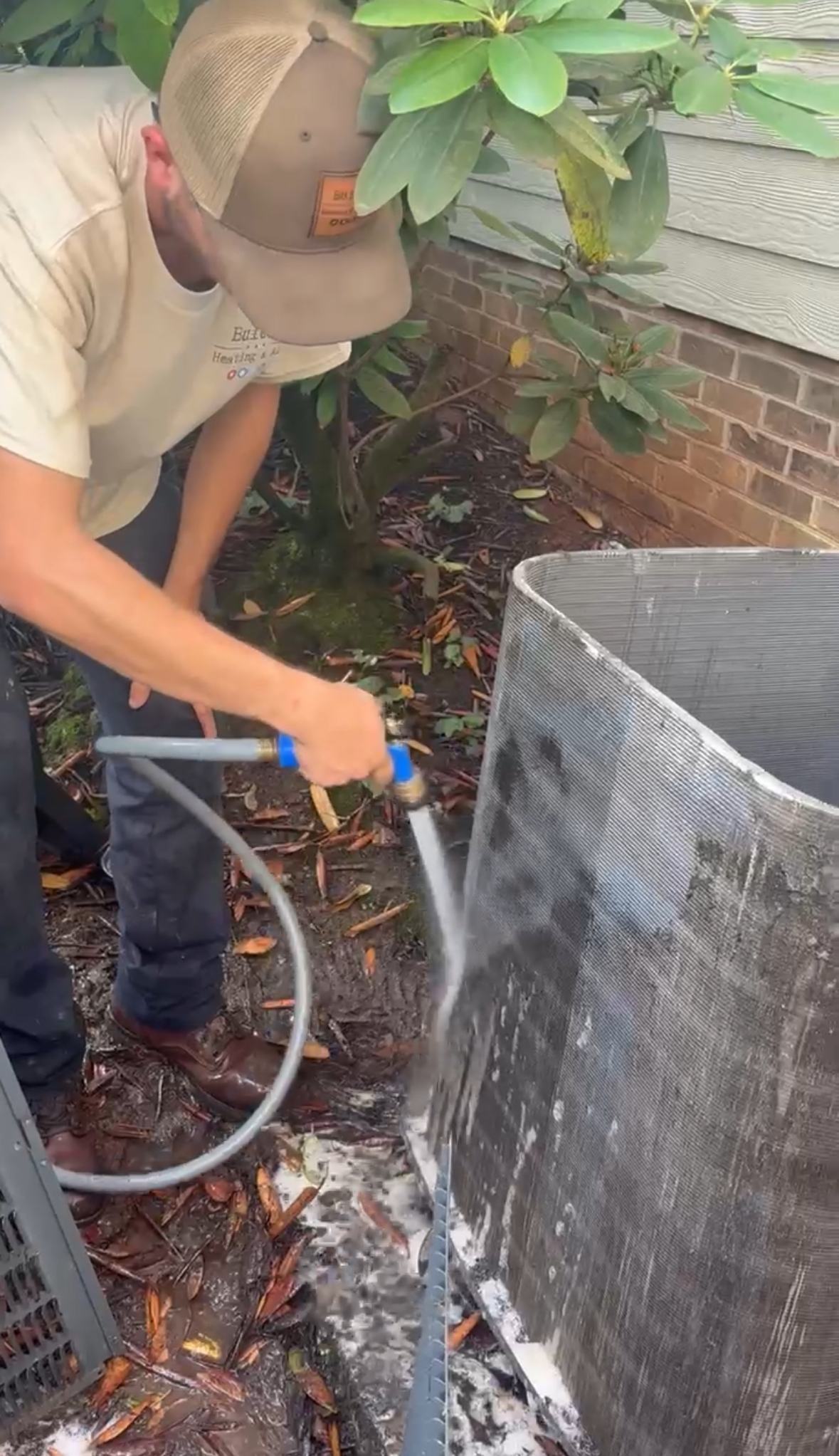 A person performs HVAC maintenance by using a hose to wash a large metal air conditioner coil outdoors, with soap and water visible on the ground.