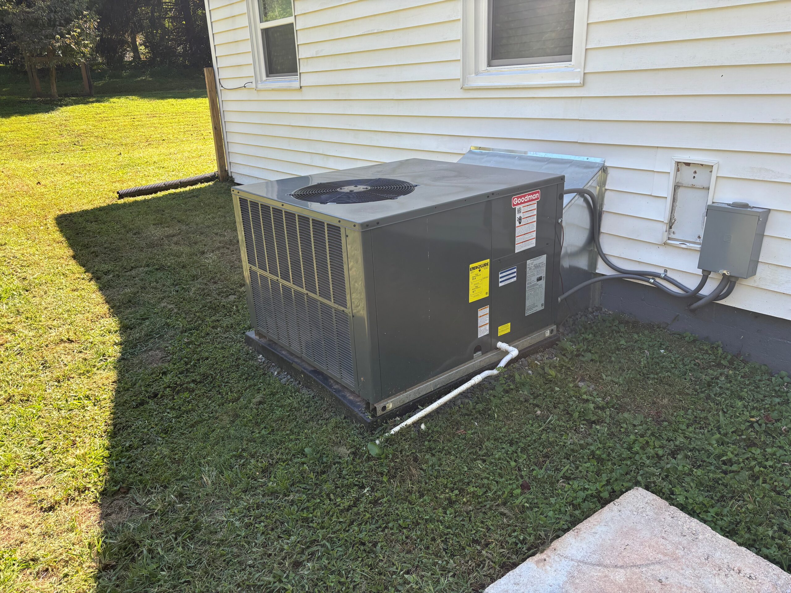 A central air conditioning unit is installed next to the exterior wall of a house, sitting on a concrete pad on the lawn.