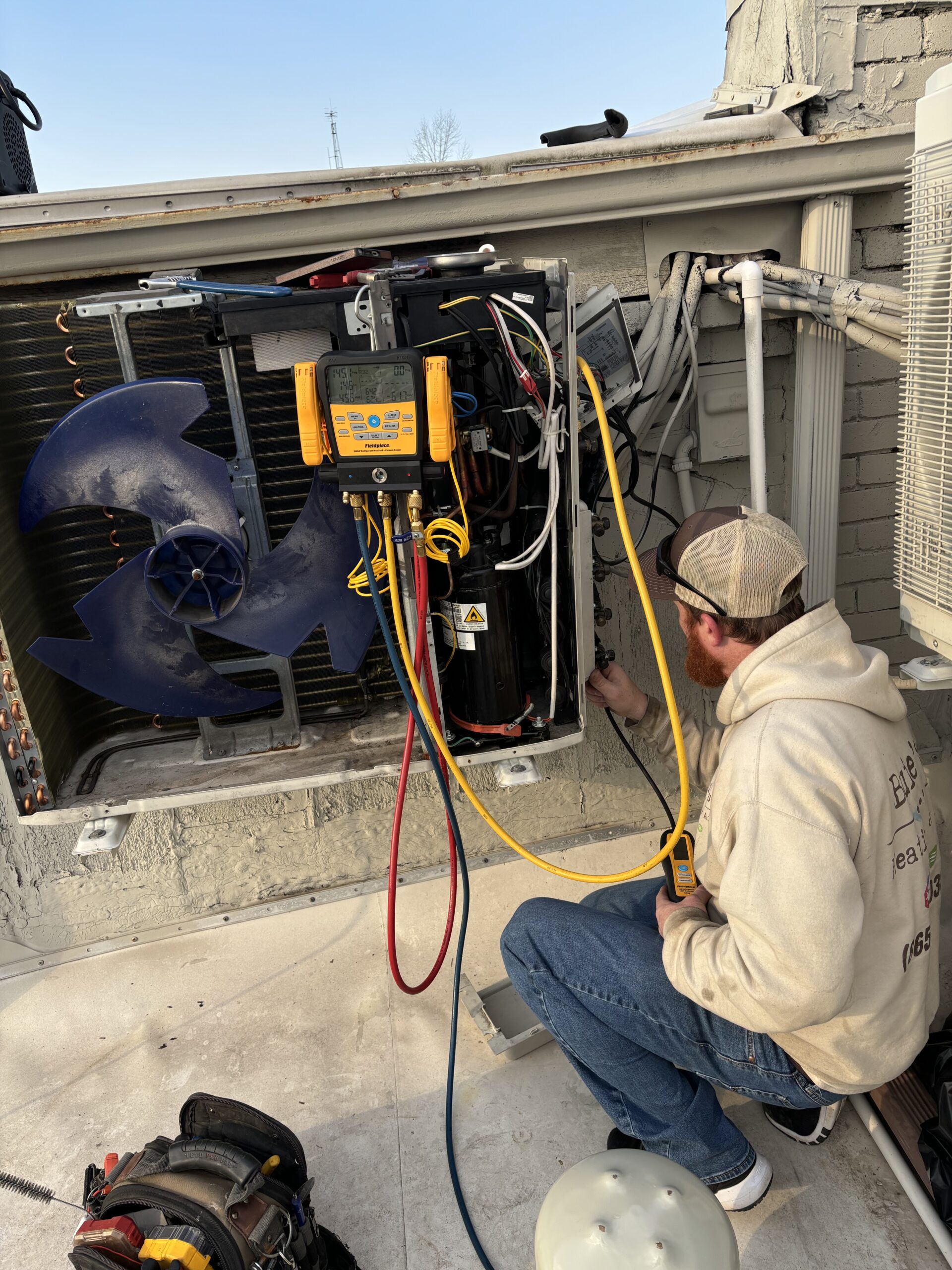 An HVAC contractor in a hoodie and cap checks an HVAC unit on a rooftop, using gauges and tools connected to the system.
