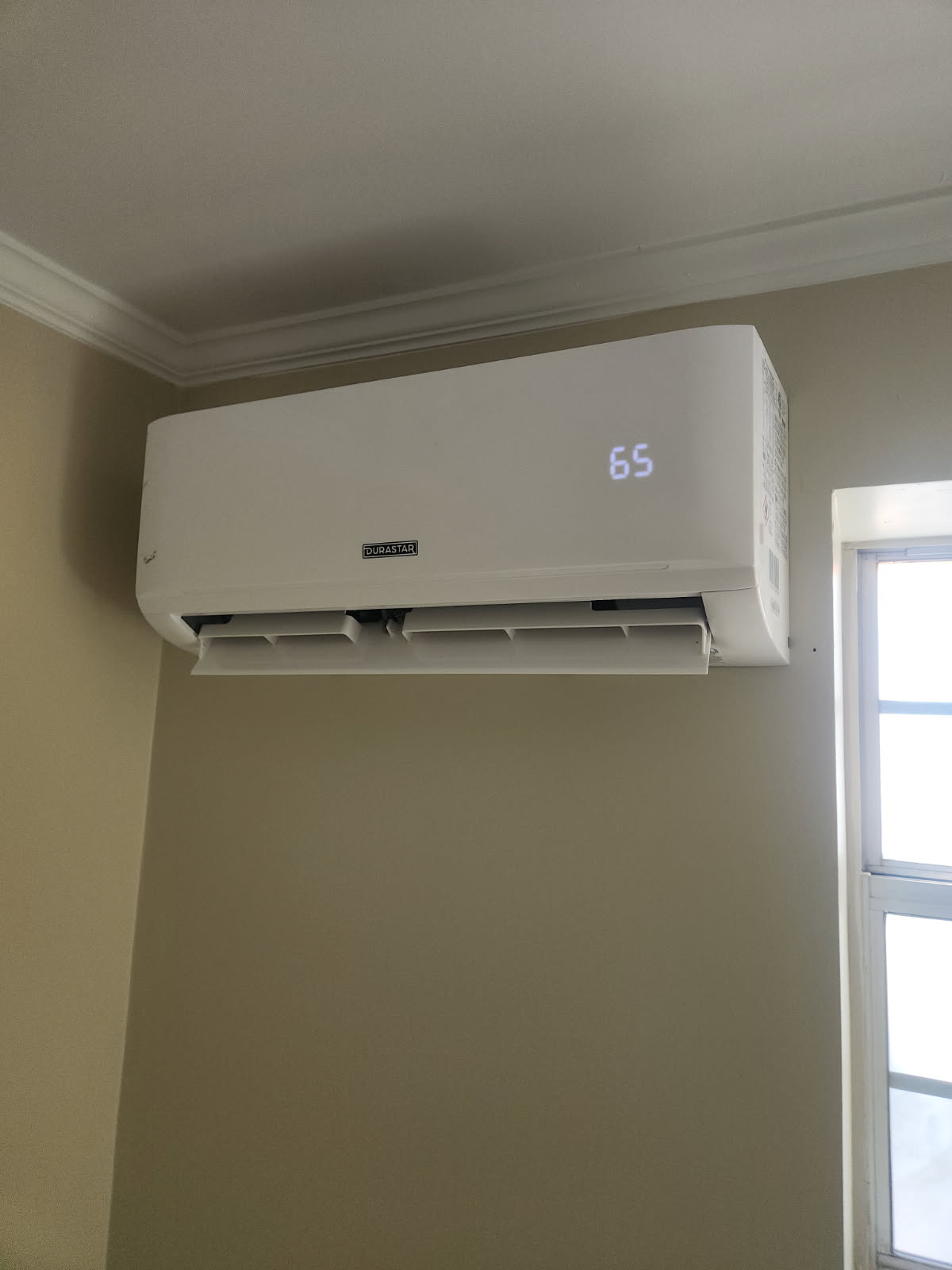A white wall-mounted air conditioner with a digital display showing 65 is installed above a window in a beige room.