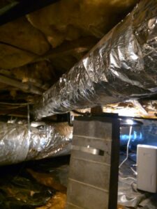 Exposed insulated air ducts run through a crawl space with insulation on the ceiling and a concrete support pillar in the foreground.