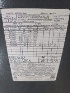 Close-up of a label on HVAC equipment showing model and serial numbers, heater kit specifications, electrical requirements, and minimum room area in square feet and square meters.