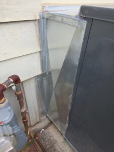 Metal ductwork connects an HVAC unit to a house exterior wall; gas meter and pipes visible to the left.