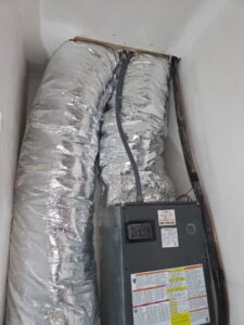 A metal HVAC unit with two large insulated air ducts and several cables is installed in a white-walled utility space.