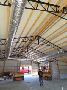 A person walks inside a large unfinished building with exposed wooden beams, metal trusses, and ductwork; construction equipment and materials are visible.