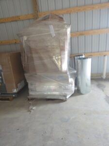 A pallet wrapped in plastic sits in a warehouse next to a palletized box and two large rolls of reflective material against a metal wall.