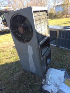A large, upright outdoor HVAC unit stands on grass, with its vent fan exposed and visible rust and weathering on its metal surface.