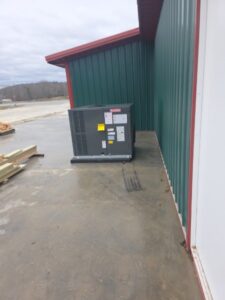 A large HVAC unit sits on a concrete slab outside a green metal building with a red roof trim on a cloudy day.