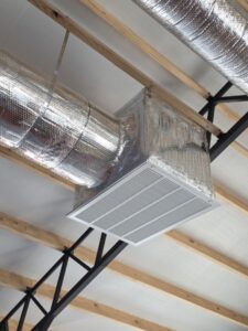 Metal ventilation duct with a rectangular air vent, wrapped in reflective insulation, mounted on the ceiling of a building with exposed wooden beams and metal supports.