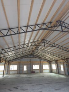 Interior of an unfinished building with exposed wooden beams, metal trusses, concrete floor, and large windows letting in daylight.