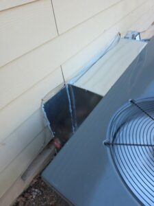 A close-up view of an outdoor air conditioning unit installed next to a house, with metal ductwork sealed along the exterior wall.