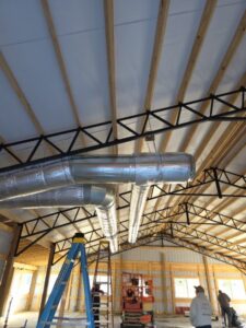 Metal air ducts are installed in the ceiling of a large unfinished building with exposed wooden beams; a blue ladder and workers are visible below.