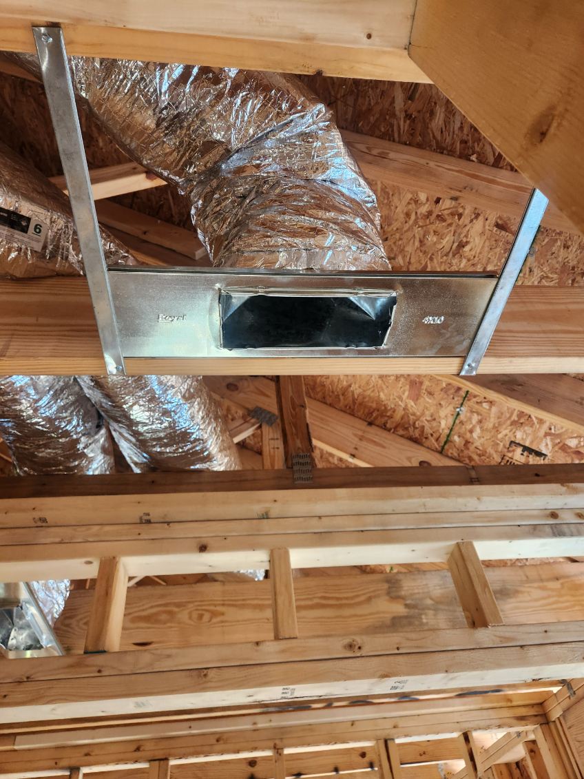 Exposed HVAC ductwork with a metal vent box installed in the ceiling of a building under construction with unfinished wooden framing.