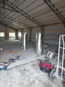 Interior of a metal-framed building under construction with tools, insulation rolls, and construction materials scattered on the concrete floor.