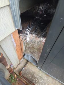 An HVAC duct covered in reflective insulation is partially exposed outside a building, next to a wooden post and metal pipes.