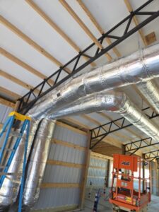 Large silver HVAC ductwork installed along the ceiling of a metal building, with a blue ladder and orange lift visible below.