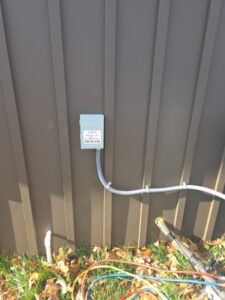 A gray electrical box is mounted on a brown metal wall with conduit running down to the ground. Fallen leaves and cables are visible on the grass below.