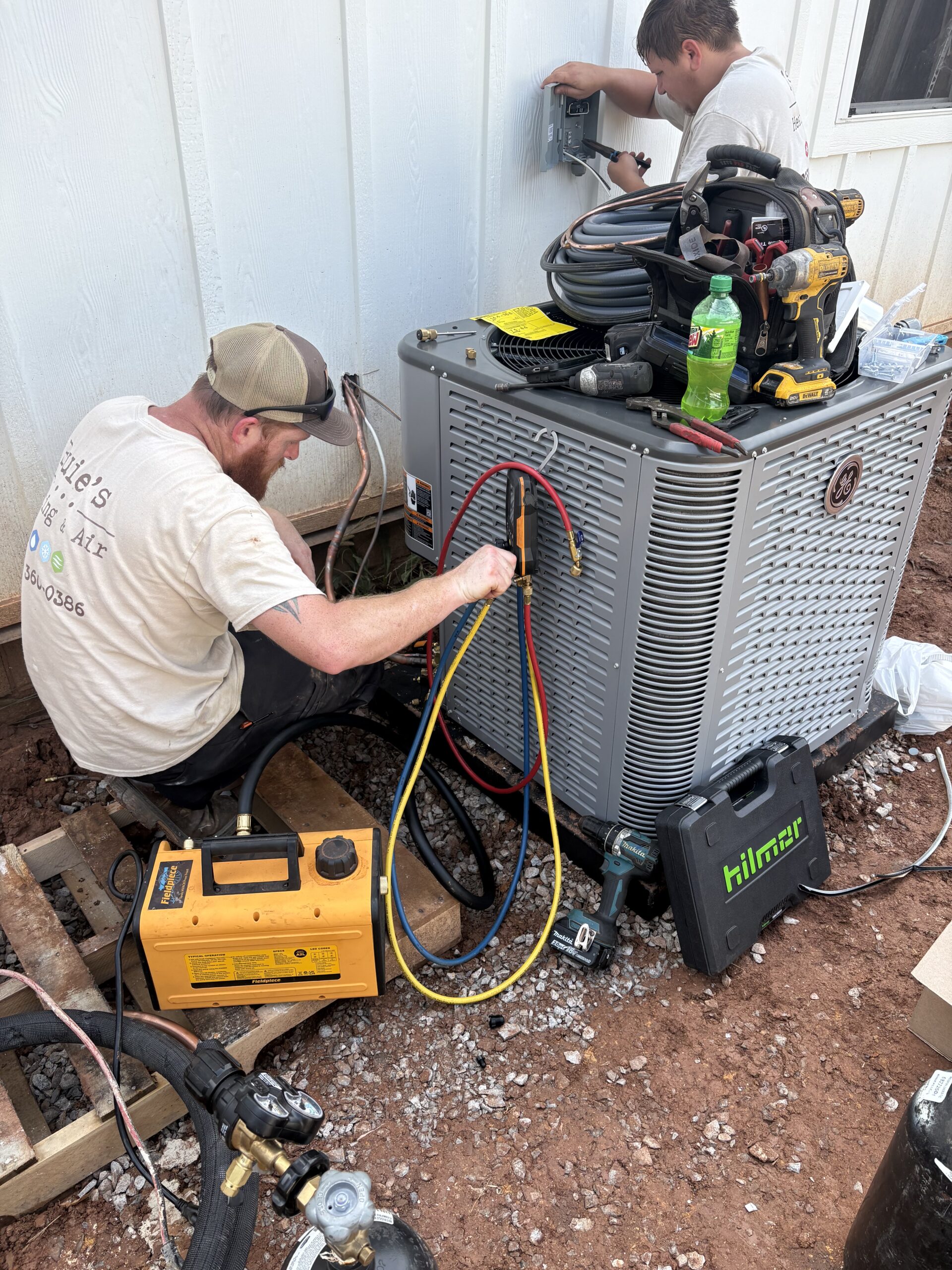 Two technicians install and service an air conditioning unit outside a building, using various tools and colored hoses connected to the equipment.