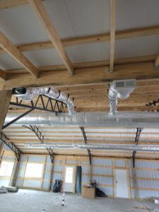 Exposed HVAC ductwork and vents installed along the wooden ceiling beams inside a large, unfinished metal building.
