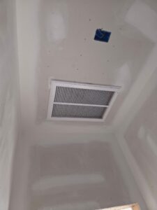 A white air vent and an uncovered electrical box are installed on an unfinished drywall ceiling with visible seams and patches.