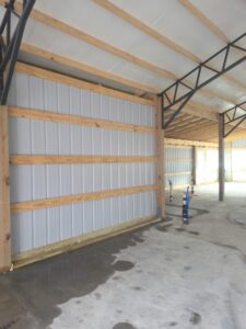 Interior of a partially constructed building with exposed wooden beams, metal siding, concrete floor, and visible plumbing pipes.
