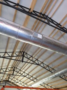 Exposed metal HVAC ductwork with an air vent mounted on a ceiling with wooden beams and black metal supports.
