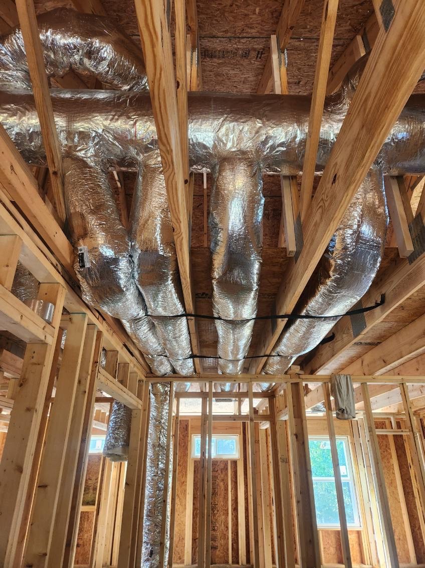 Exposed ductwork wrapped in insulation runs through the unfinished wooden framing of a house during new construction and additions HVAC installation.